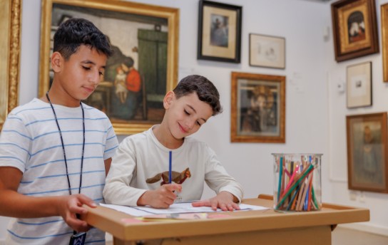 Twee kinderen tekenen in een museum, omringd door schilderijen. (vergroot foto)
